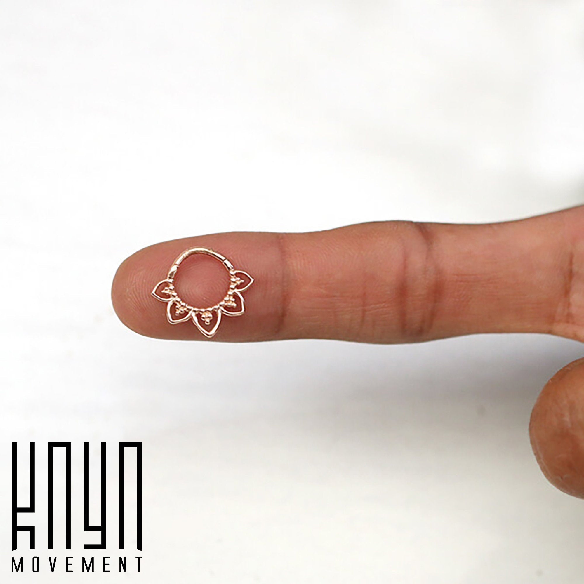 A close-up of a person's finger holding a small, intricate ring. The MAWAR Lotus Clicker Septum Ring, crafted from surgical steel with a rose gold PVD coating, features a delicate floral design. The image has a white background and the text "Kaya Movement" is displayed in the lower left corner.